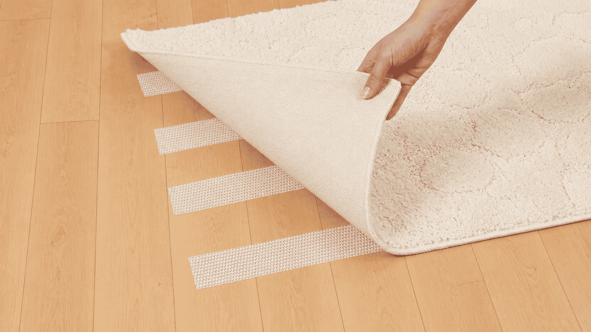 hand lifting a light cream rug to reveal non slip adhesive strips placed across a smooth maple wood floor