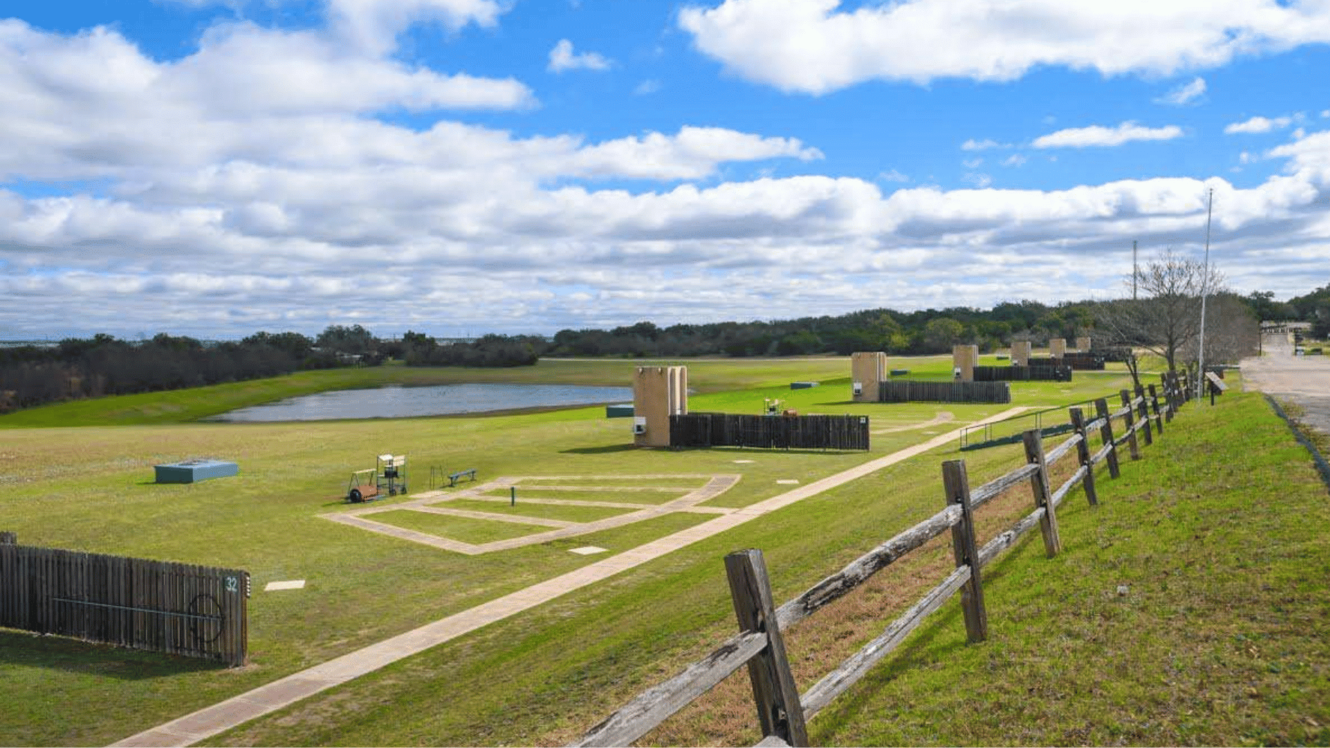 green open landscape with walking paths, wooden fences, water pond, and cloudy blue sky in a park area