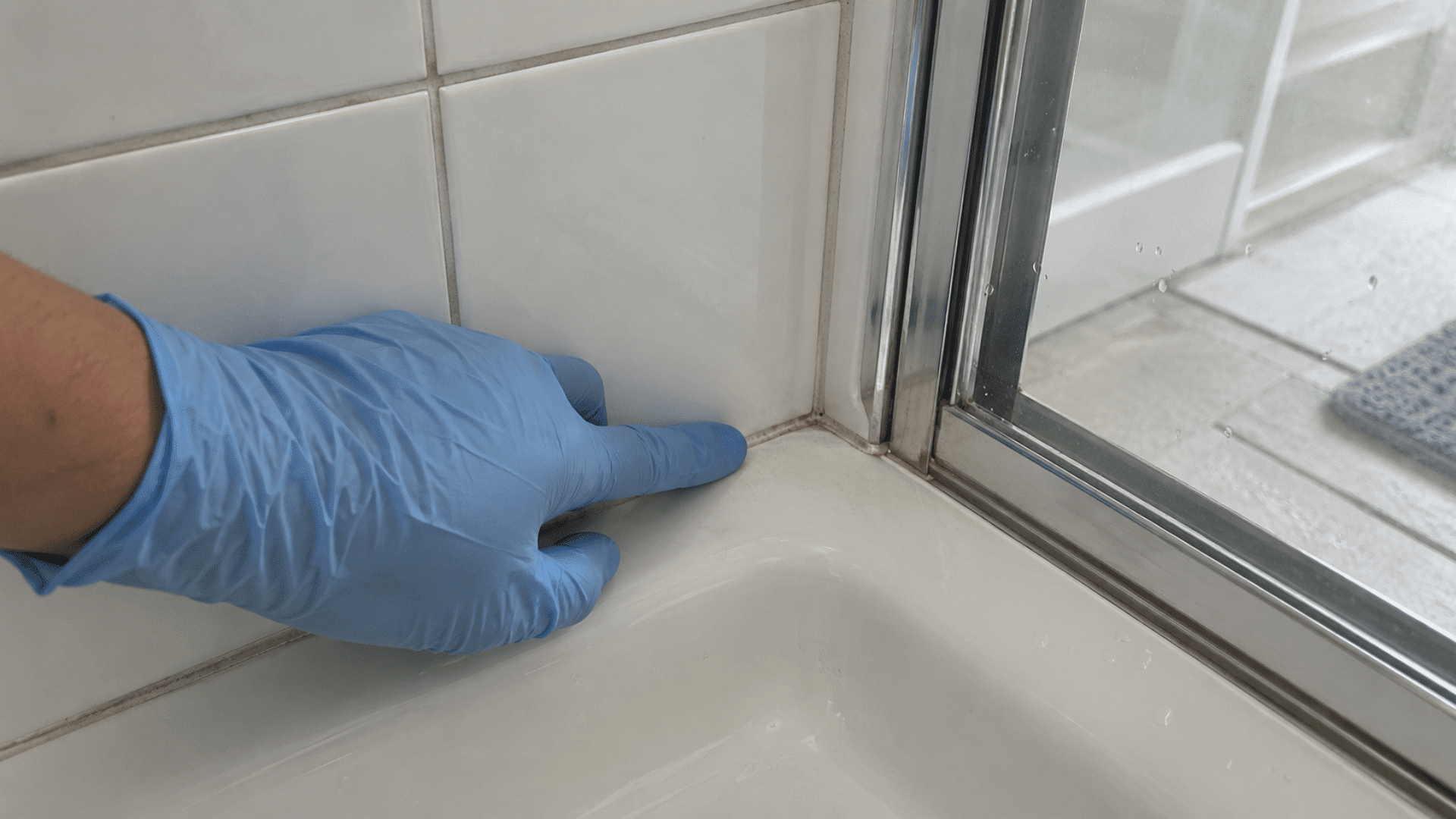 blue-gloved hand checking the shower corner and grout before cleaning