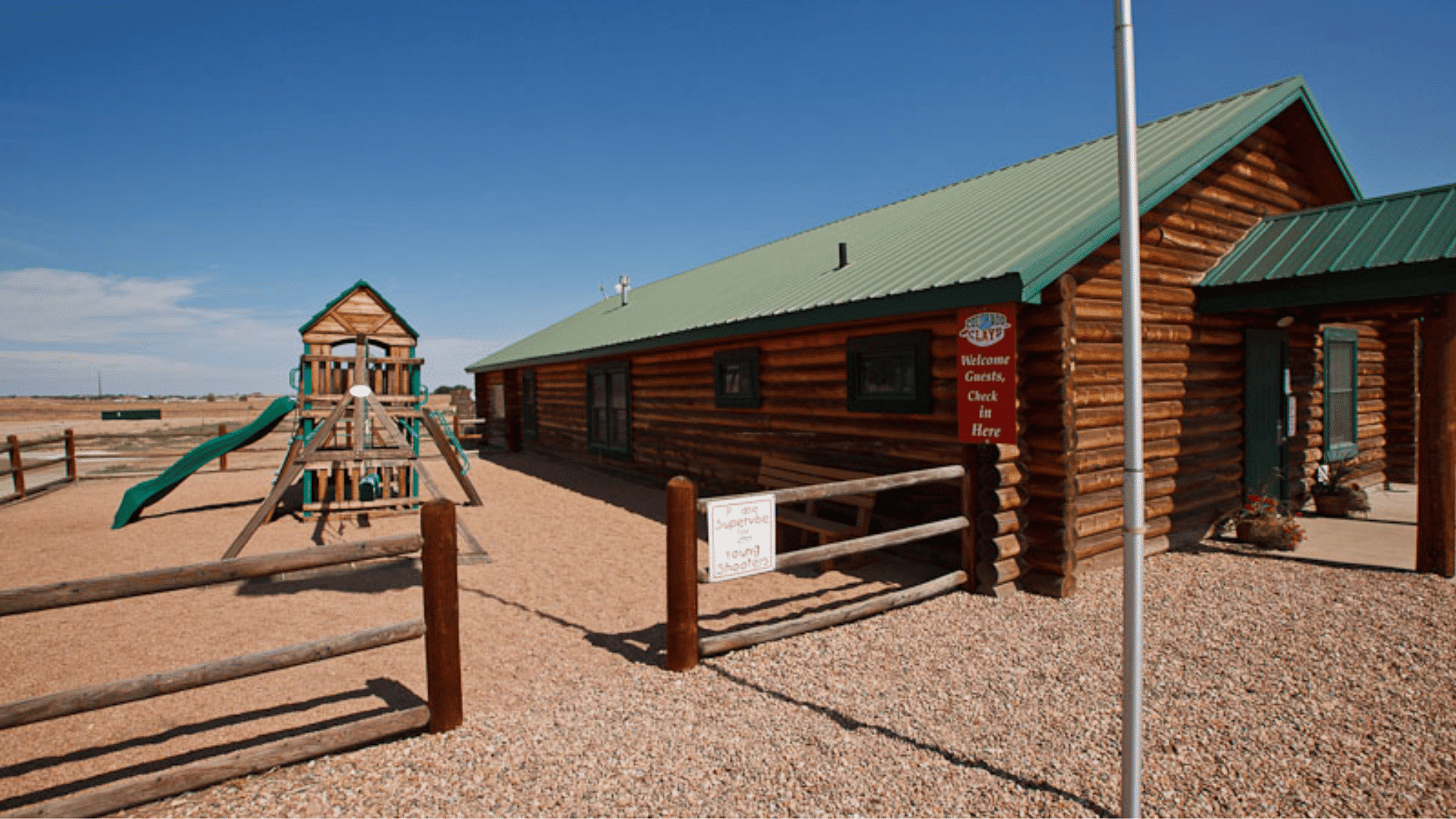 log cabin building with playground area, gravel yard, wooden fence, and clear blue sky outdoors