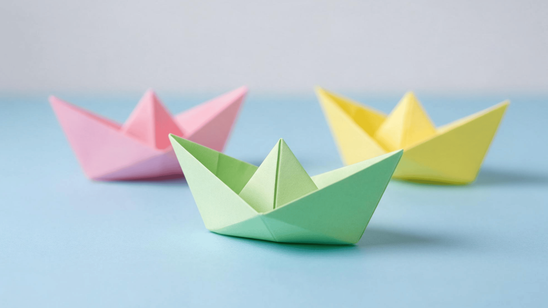 three pastel origami paper boats, green, pink, and yellow, on a blue surface