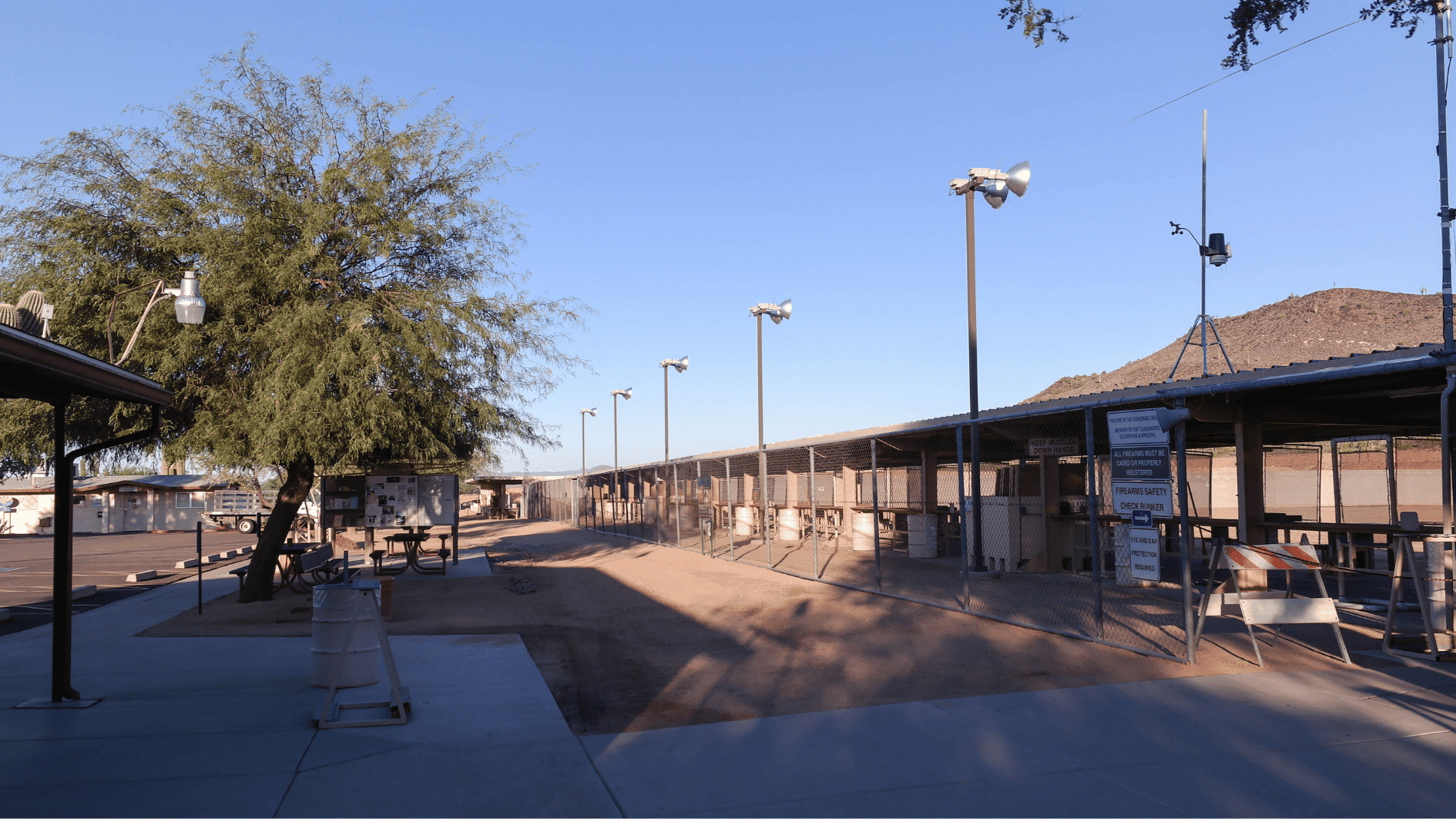 outdoor shooting range with covered shooting lanes, safety signs, fencing, and desert landscape background