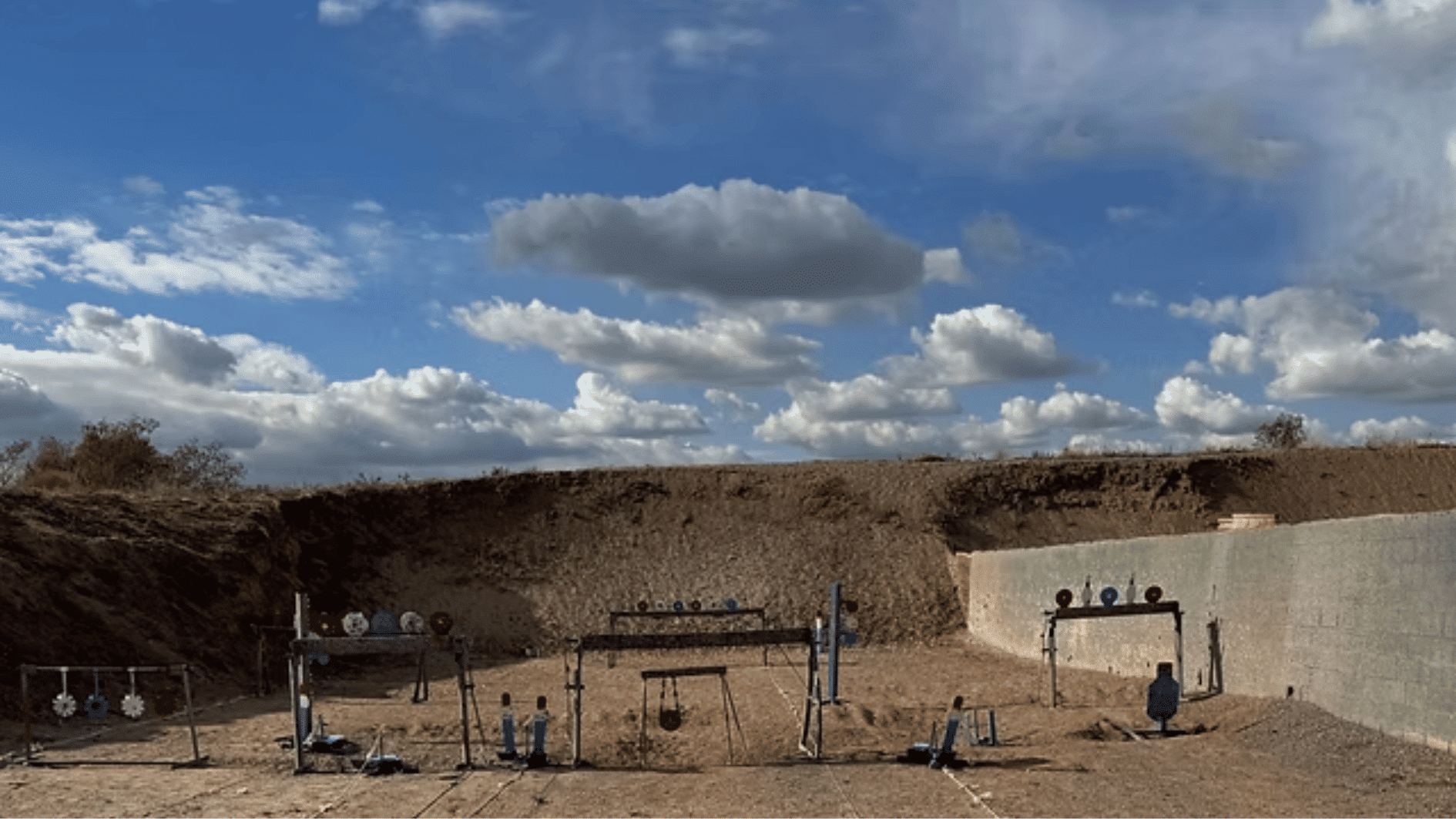 outdoor shooting range with target stands, dirt berm, concrete wall, and cloudy blue sky background