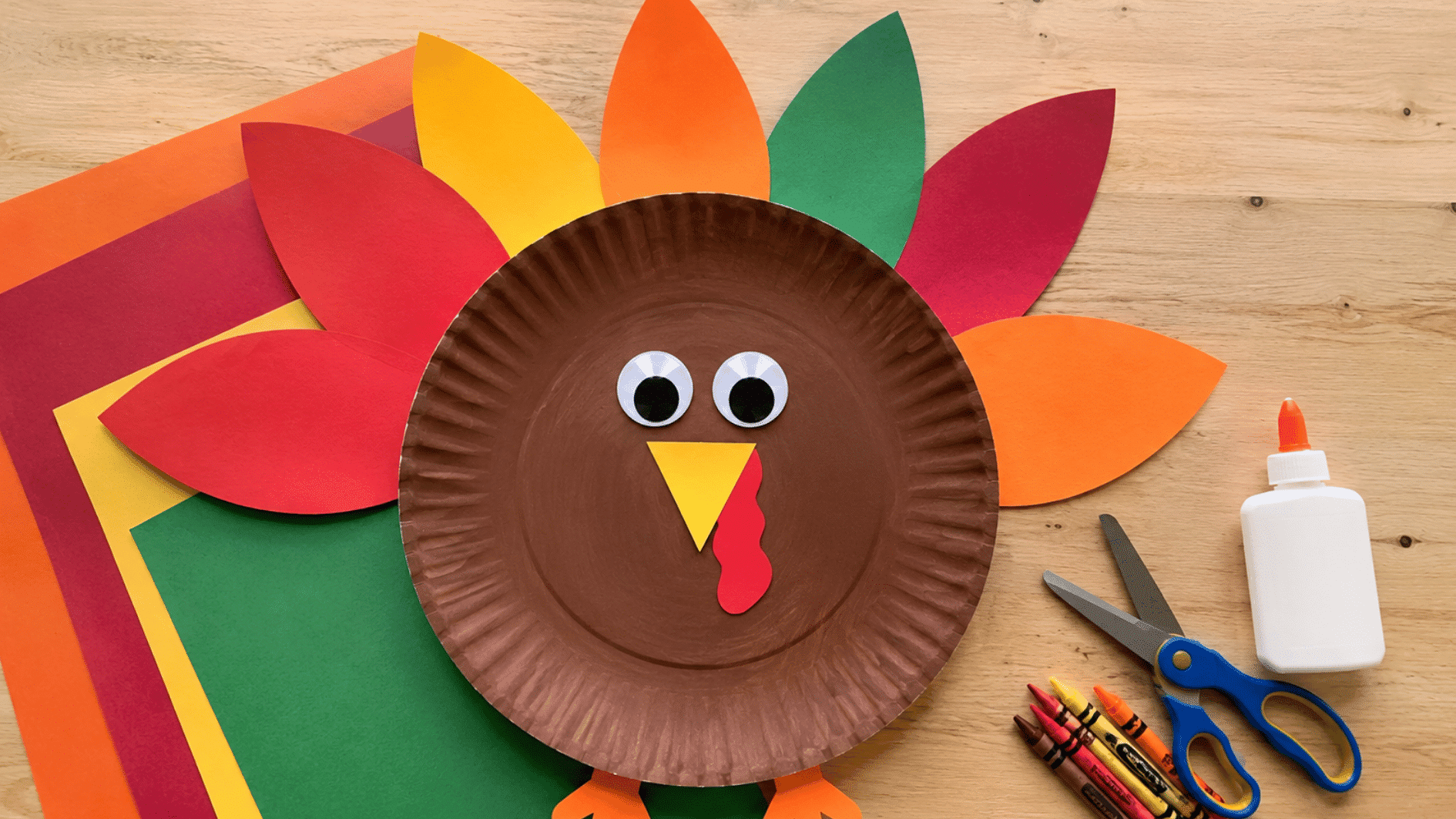 paper plate turkey craft with colorful paper feathers, beak, wattle, and feet on a wooden table
