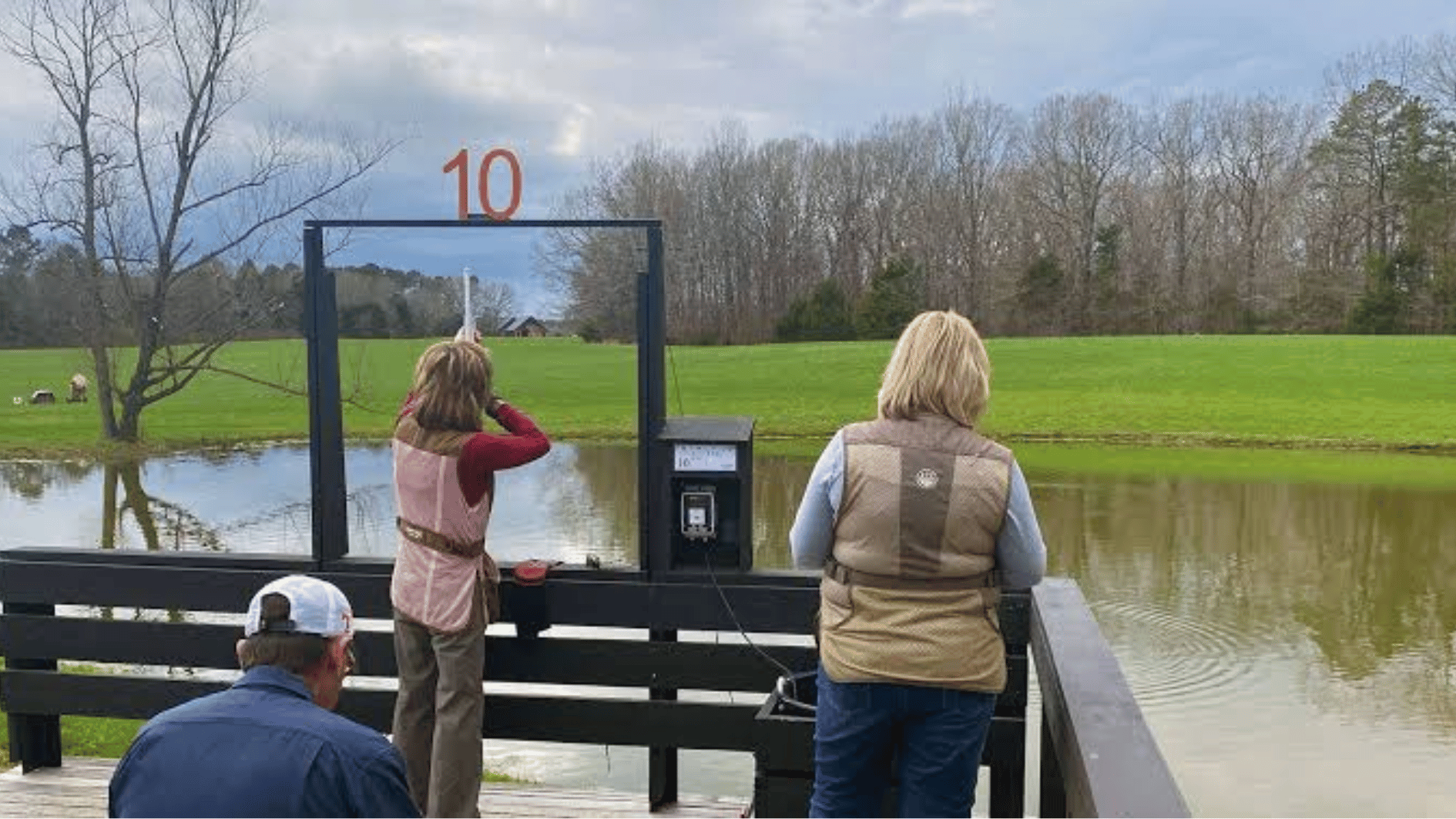 people at an outdoor clay shooting station beside a pond with green field and trees in background