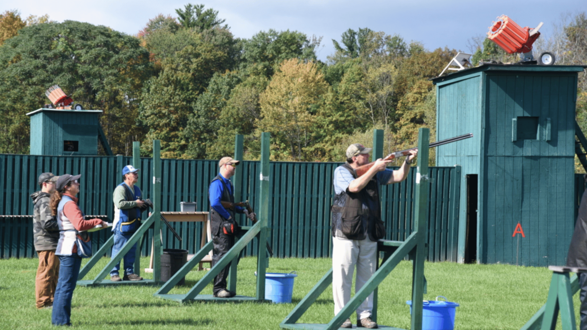 people practicing clay shooting at an outdoor range with target launchers, green fields, and wooded background