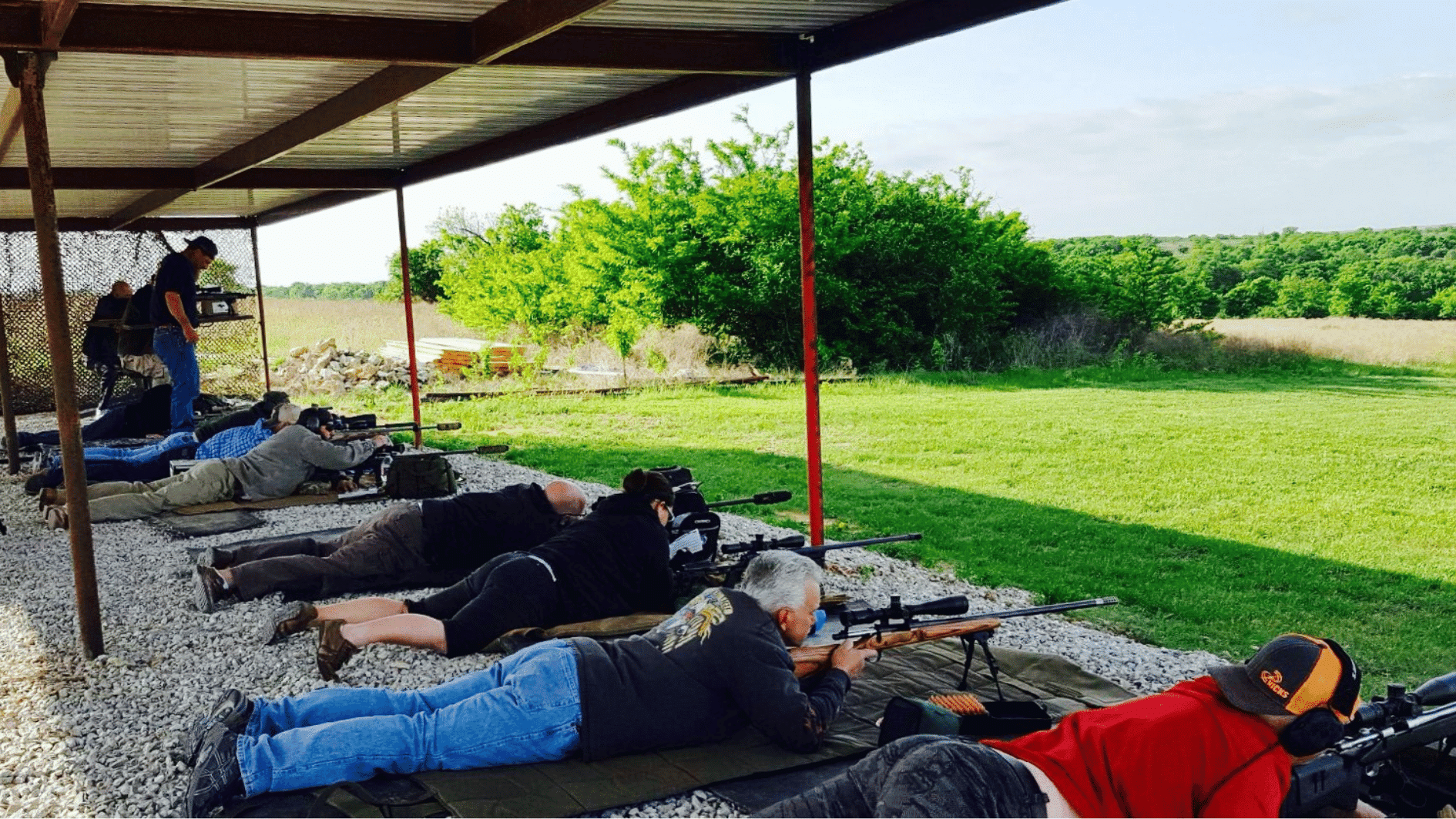 people practicing target shooting at an outdoor range under a covered shelter with green field background
