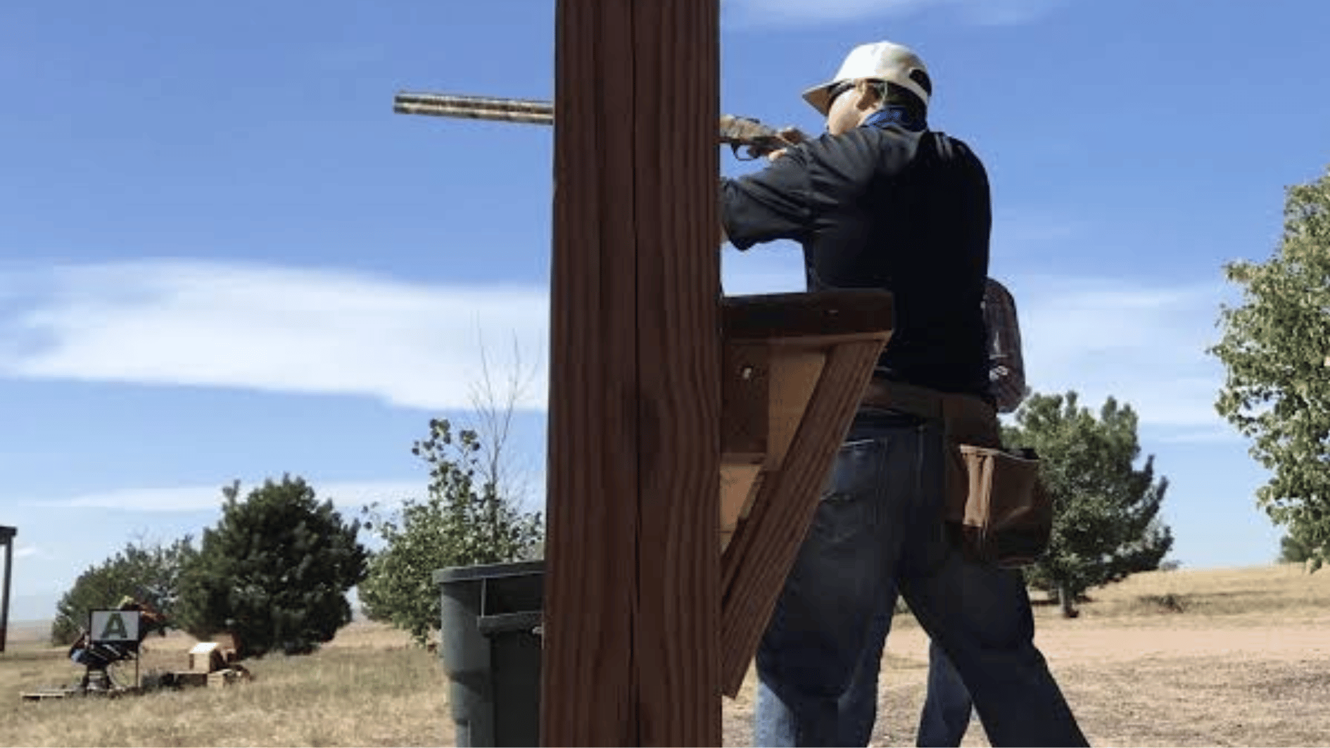 person practicing shooting at an outdoor range under a wooden shelter with blue sky background
